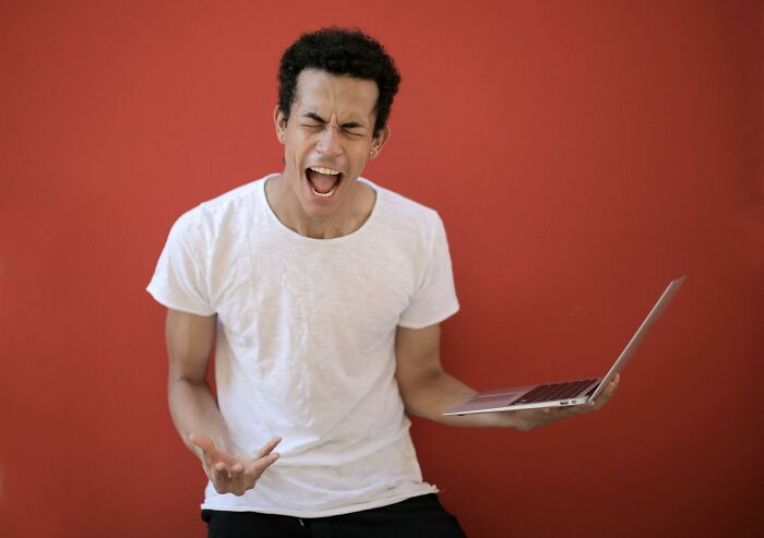 Young man frustrated holding laptop, reacting to tricky questions challenge against solid red background.