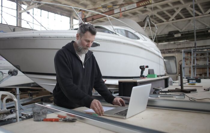 Man working on laptop and measuring tools inside a workshop with a luxury yacht, highlighting jobs for ultra-wealthy people.