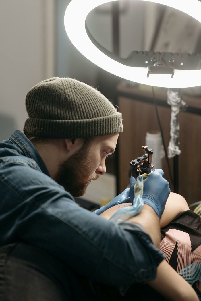 Tattoo artist wearing gloves and a beanie focused on tattooing a client’s arm under bright ring light in a studio setting