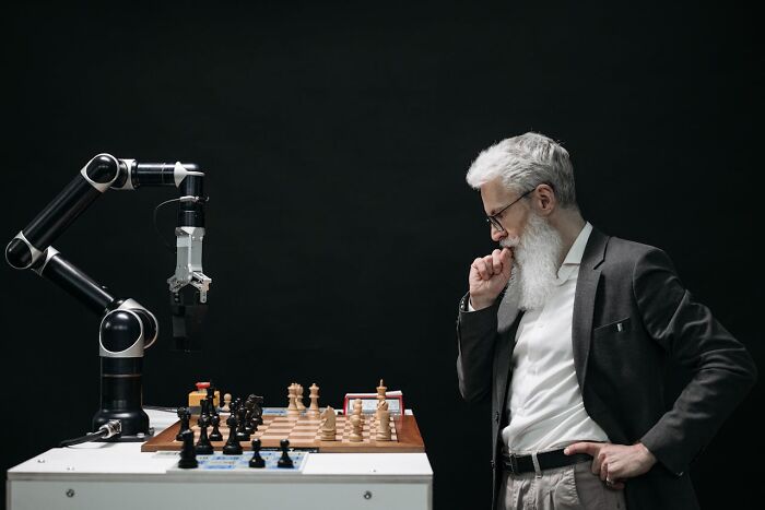 Elderly man with white beard playing chess against robotic arm, illustrating jobs for ultra-wealthy people concept.
