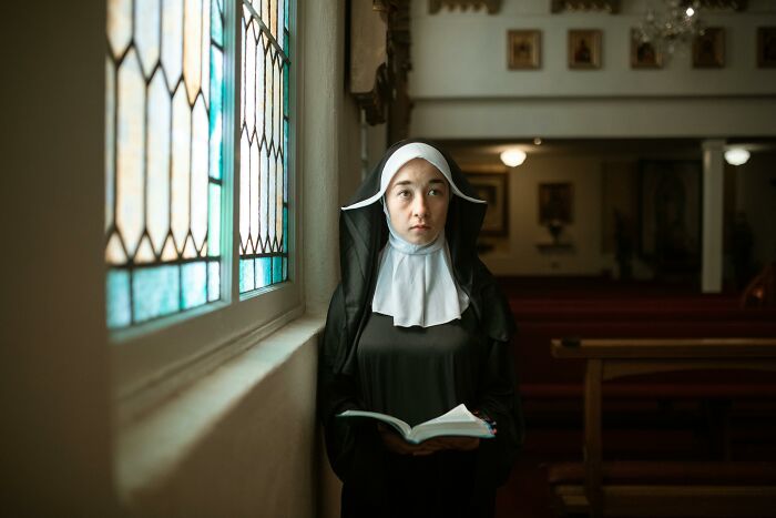 Young woman dressed as a nun standing by stained glass, holding a book, symbolizing sweet facade hiding manipulation and betrayal.
