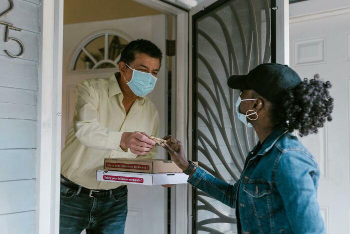 Man wearing a mask handing over pizza boxes to a masked delivery person representing overhyped products buying trend.