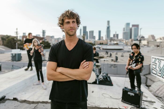 Man with crossed arms on rooftop while people share sounds in an extremely angry band rehearsal with city skyline background