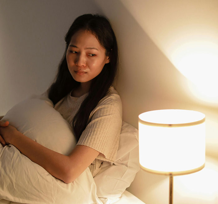 Hurt working mom sitting alone in bed holding a pillow, looking upset and tearful beside a lit lamp.
