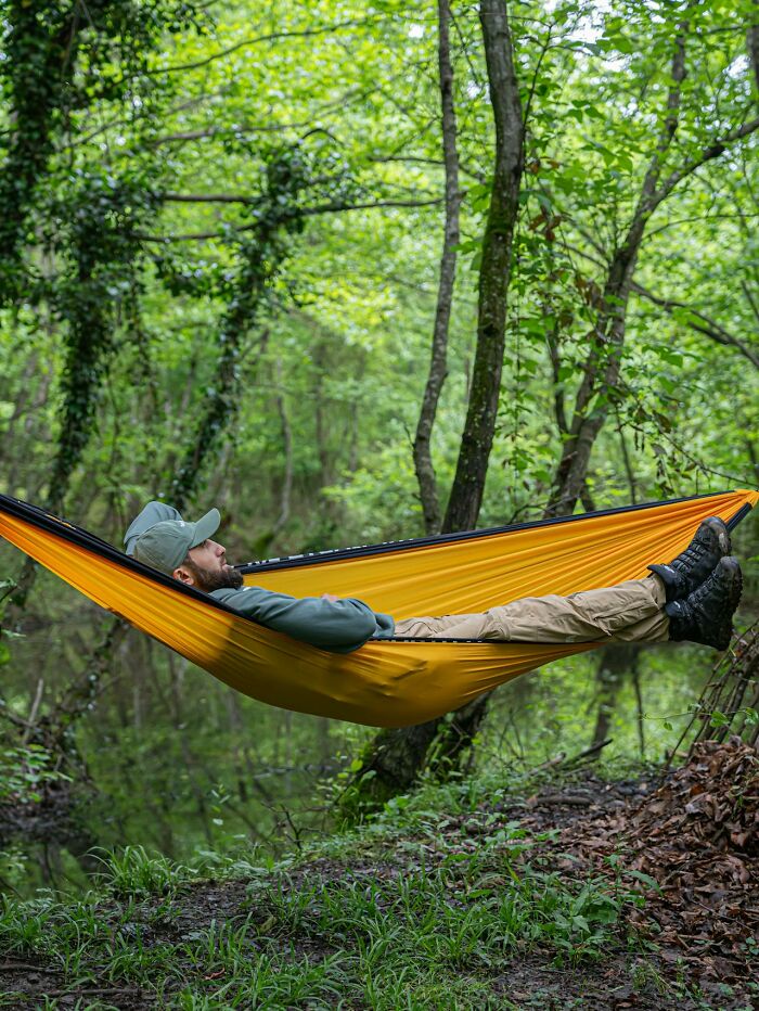 Man relaxing alone in a yellow hammock in the woods surrounded by green trees and foliage, embodying solitude in nature.