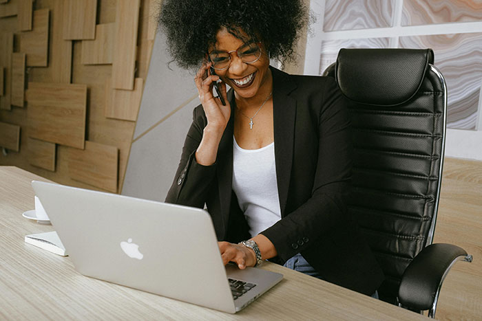 Smiling working mom in glasses talking on phone while using laptop at office, representing hurt working mom and ignorant husband theme