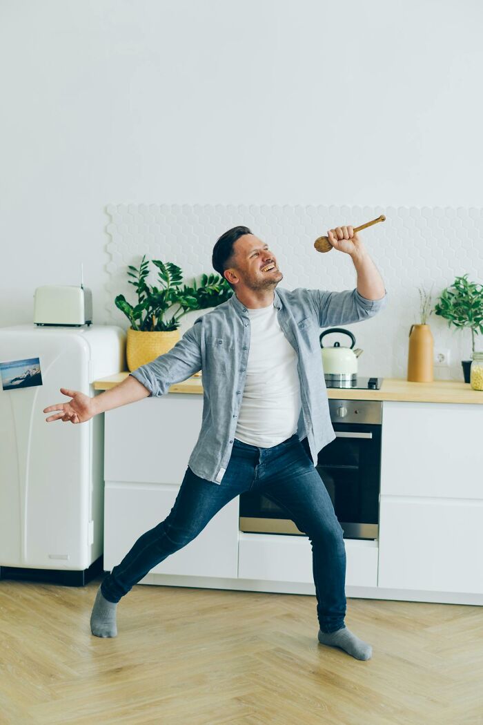 Man in casual clothes singing in kitchen using wooden spoon, showing carefree and dumb behavior at home.