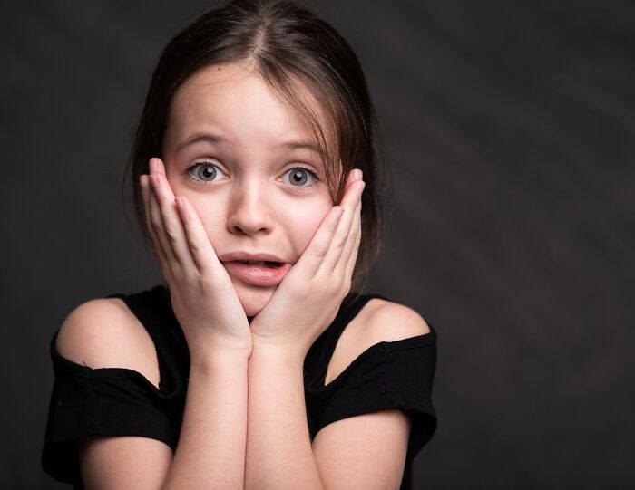Young girl looking scared and alone, holding her face with both hands against a dark background.