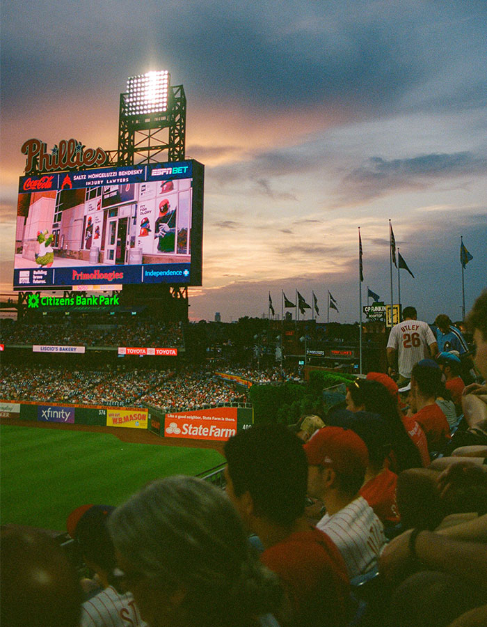 Crowd watching a Phillies baseball game at Citizens Bank Park during sunset with scoreboard displaying Phillies mascot.