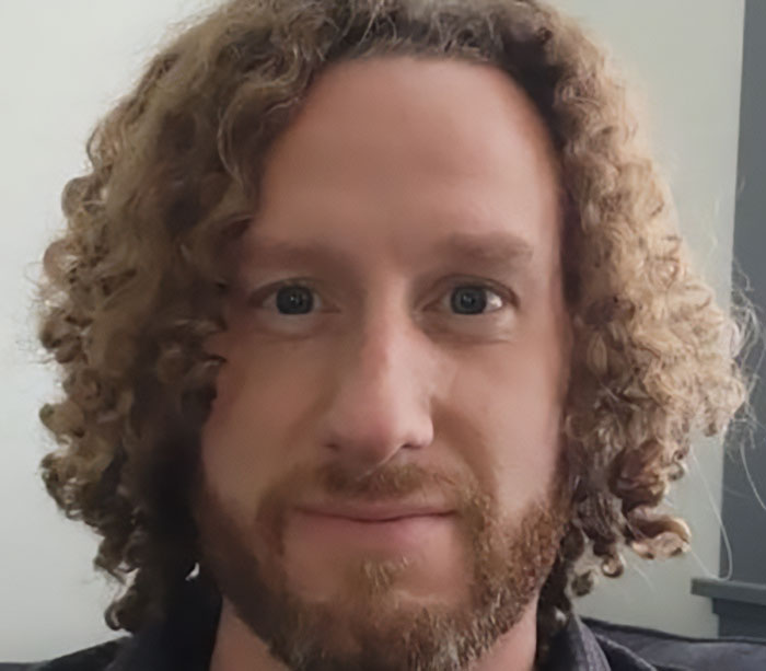 Bearded man with curly hair looking back in crowd during Charlie Kirk attack, spotted cheering with a neutral expression.