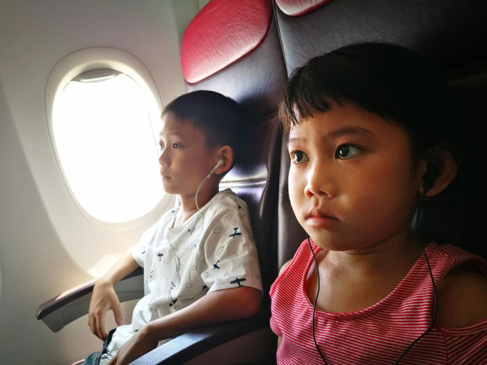 Two children wearing earphones seated on airplane seats, with a window visible beside them during the flight.