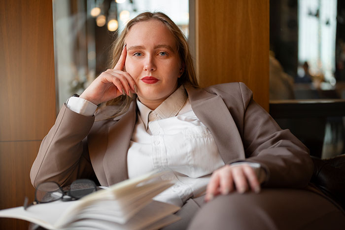 Confident woman manager in a beige suit, sitting thoughtfully with an open book, representing new manager and client fitness concerns.