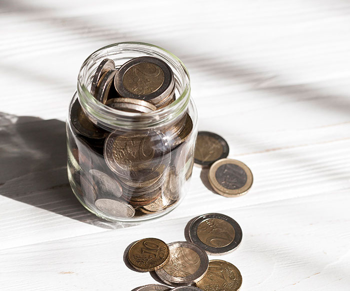 Glass jar filled with coins and more coins scattered on a white wooden surface symbolizing money habits.