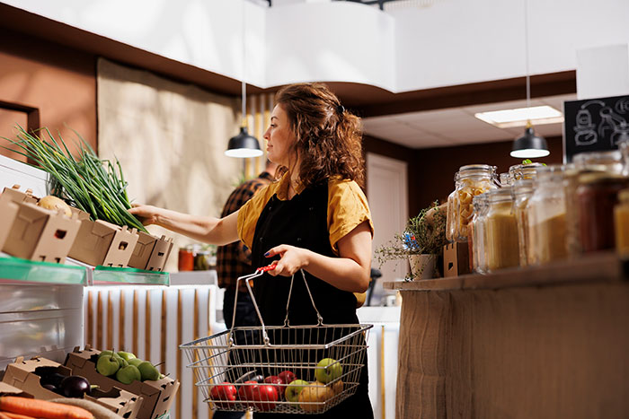 Woman with a shopping basket picking fresh produce in a grocery store highlighting habits people who used to be poor keep.