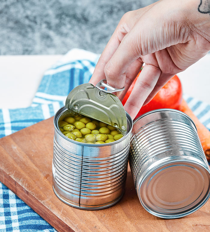 Hand opening a tin can of peas on a wooden board, illustrating habits people who used to be poor can't get rid of.