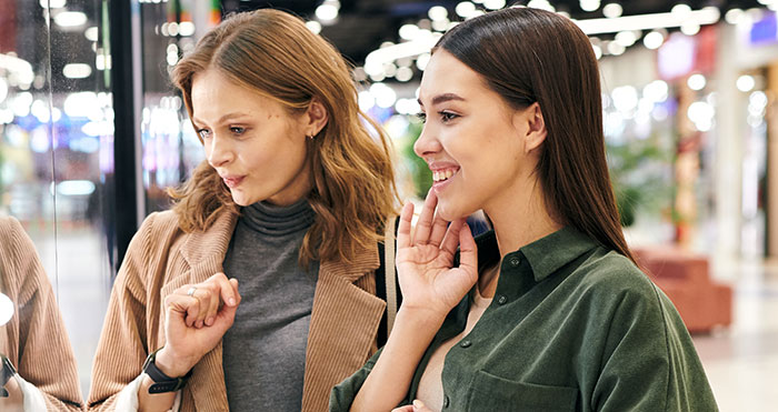 Two women smiling and discussing habits people who used to be poor can't get rid of while shopping in a mall.
