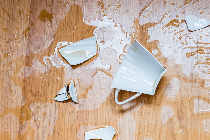 Broken white cup and spilled liquid on wooden floor illustrating habits people who used to be poor can't get rid of.