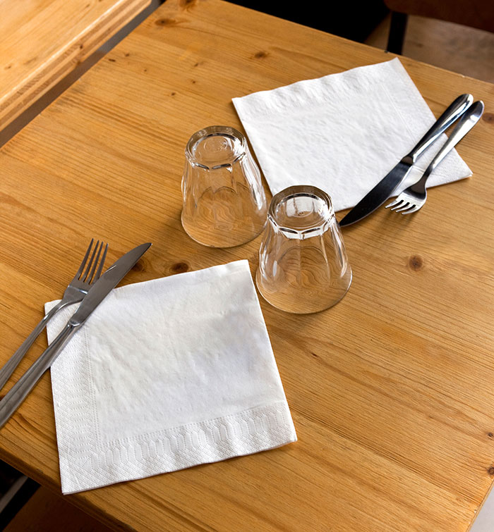 Two place settings with glasses, knives, forks, and napkins on a wooden table showing habits people who used to be poor keep.