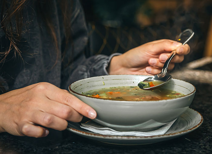 Person holding a spoon with vegetable soup in a bowl, illustrating habits people who used to be poor can't get rid of.