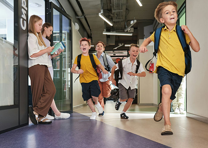Kids running down a school hallway with backpacks, while two adults stand nearby reading a book, showing product manipulation.