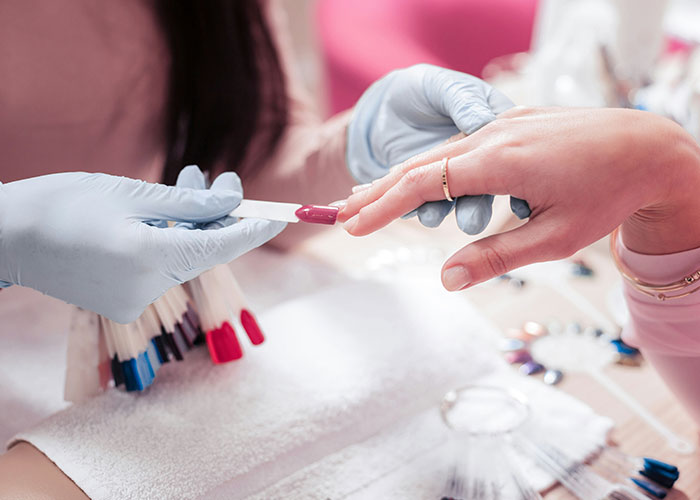 Manicurist wearing gloves applying nail polish to a person's fingernail, showcasing product in a salon setting.