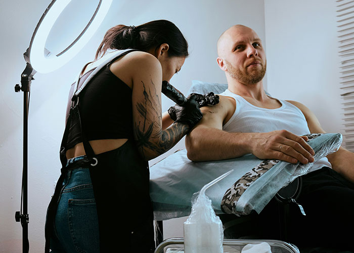 Tattoo artist manipulating person’s skin while client receives product in a professional studio setting.