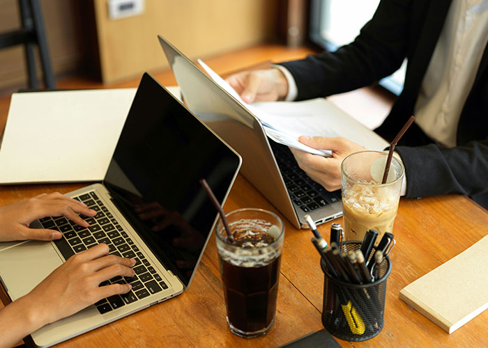Two people working on laptops at a wooden table, reviewing documents and drinking iced beverages, illustrating job descriptions.