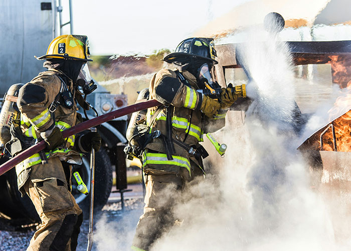 Two firefighters in full gear using a hose to extinguish a blazing fire with smoke and flames visible.