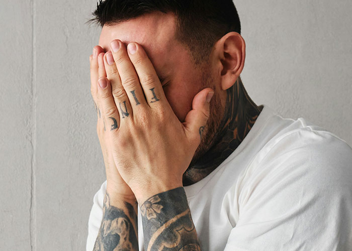 Man with tattoos covering face with hands, expressing frustration or stress related to job descriptions.