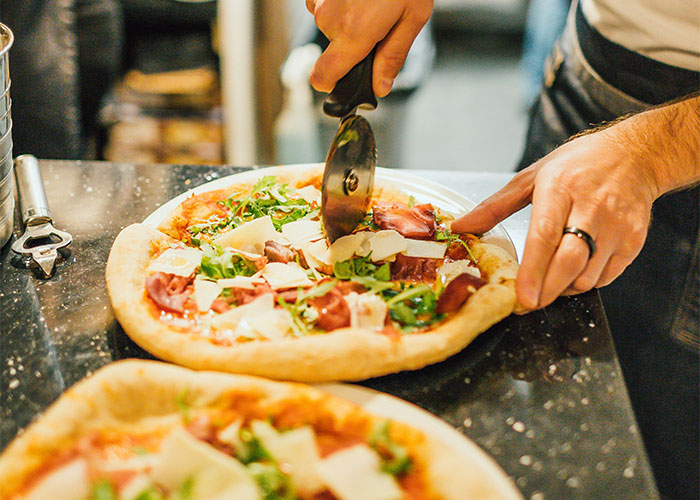 Person manipulating pizza cutter to slice freshly made product on countertop in a busy kitchen setting.