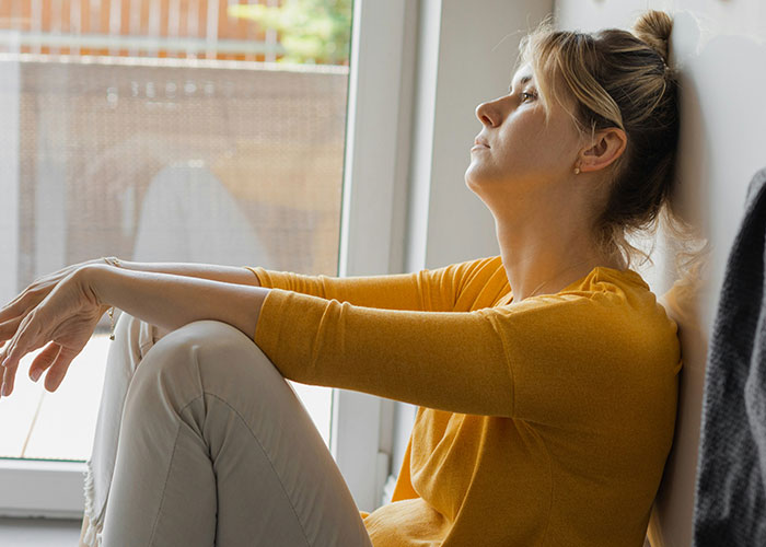 Young woman in a yellow sweater sitting by a window looking thoughtful, illustrating funny job descriptions about product manipulation.
