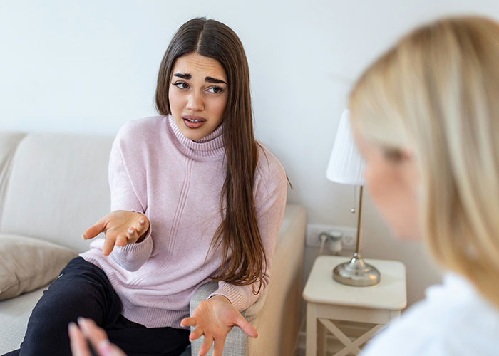 Young woman in a pink sweater explaining and gesturing while talking to another person in a casual indoor setting.