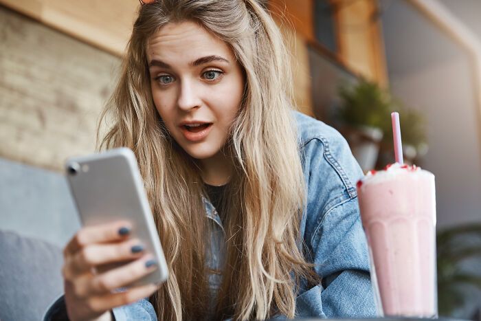 Young woman looking surprised at phone screen while sitting in cafe, relating to tests for dating red flags.