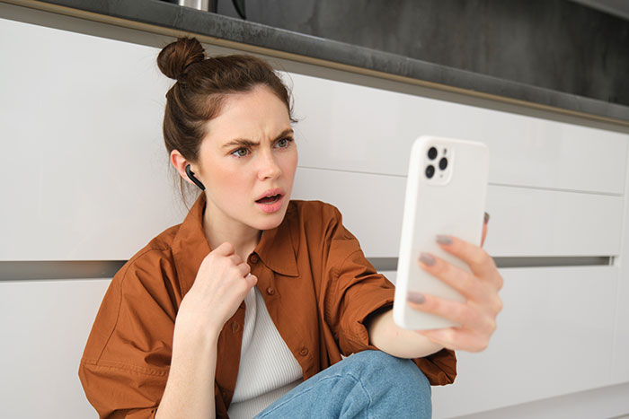 Young woman sitting on floor, looking shocked while holding smartphone, illustrating friend house sit dog conflict story.
