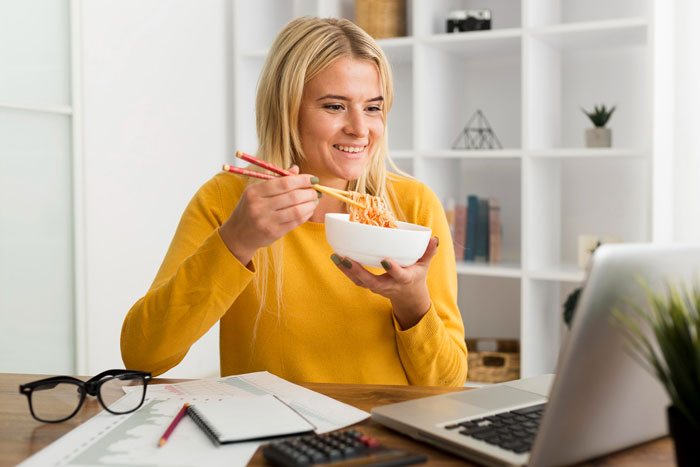 Young woman in yellow sweater eating noodles while working on a laptop, illustrating a serial food thief with peanut allergy story.