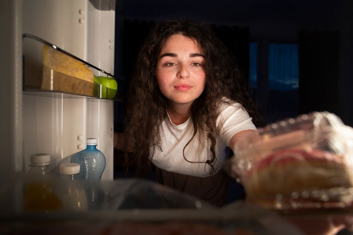 Young woman reaching into fridge at night, illustrating a serial food thief with peanut allergy stealing roommate&rsquo;s dinner.