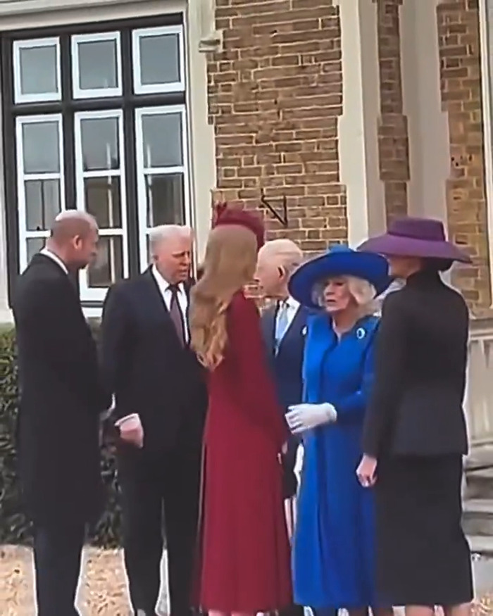 Camilla in a blue outfit making a cold gesture towards Kate and others outside a brick building during an event.