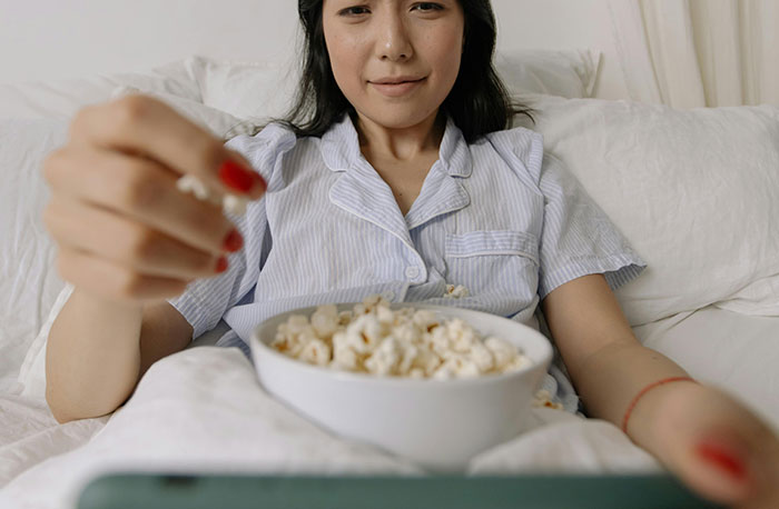 Young woman in pajamas eating popcorn in bed while watching shows about medical diagnoses on screen.
