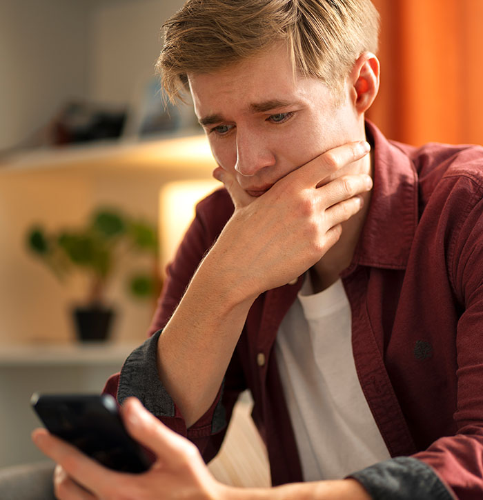 Young man in a red shirt reacts with fear and shock while reading scary real-life stories on his smartphone.
