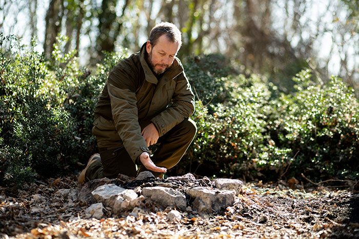Man in a green jacket kneeling in forest, examining ground with rocks, evoking scary real-life stories atmosphere.