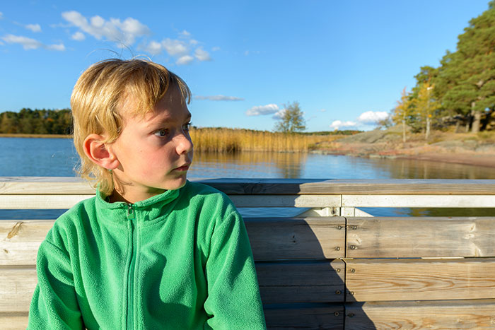 Young boy in a green jacket sitting by a lake, looking uneasy, reflecting scary real-life stories people can’t tell.