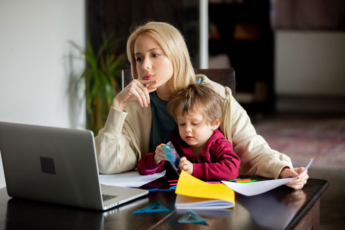 Stepmom looking tired and overwhelmed while babysitting her young stepkid at a table with laptop and papers.