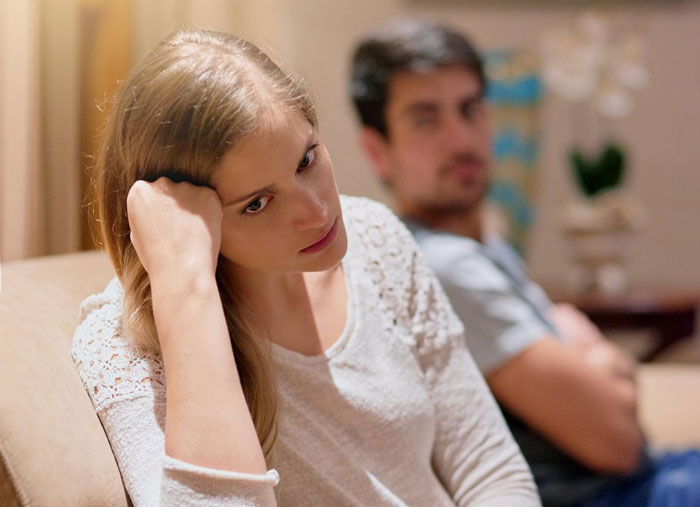 Stepmom looking exhausted and stressed while sitting on a couch with a man blurred in the background.