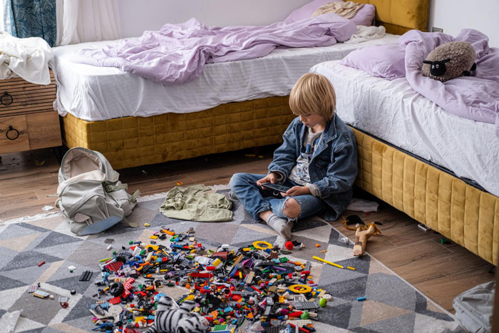 Young boy sitting on a messy bedroom floor with toys scattered, illustrating the teen creates hygiene checklist for bro before school.
