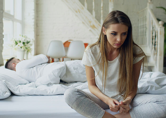 Upset wife sitting on bed looking away while man lies down, reflecting tension over baby name and first girlfriend. Upset wife sitting on bed looking away while man lies down, reflecting tension over baby name and first girlfriend.