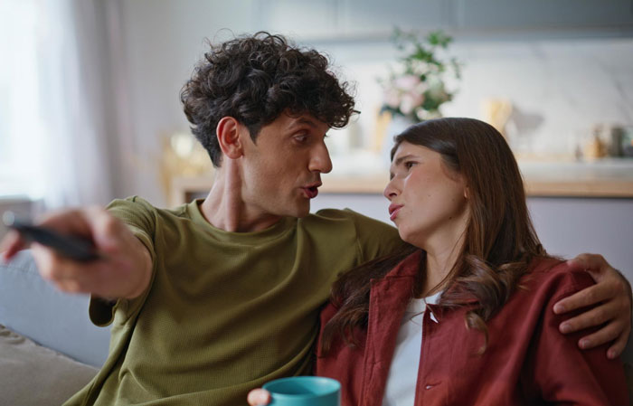 Young man refusing to watch autistic friend's show recommendation while sitting closely on a couch at home.
