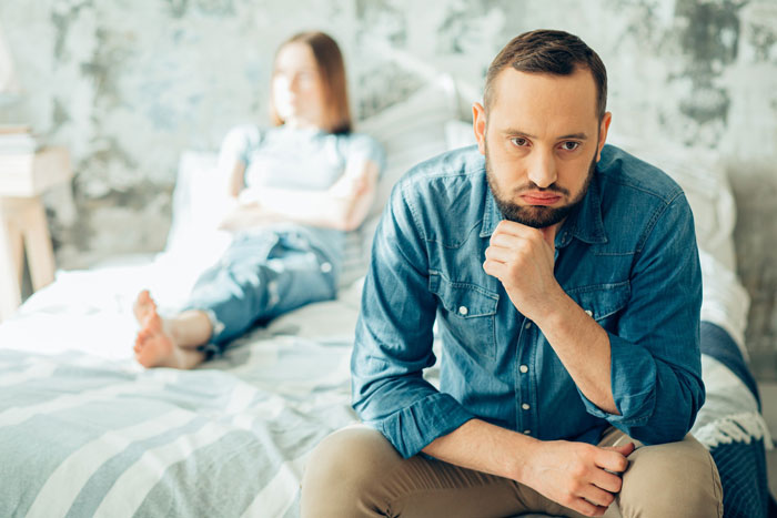 Man looking regretful sitting on bed with partner blurred in background, reflecting on marriage ruined by online affair.