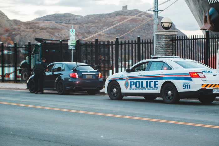 Police officer talking to driver of black car during traffic stop on roadside with police car nearby at dusk.