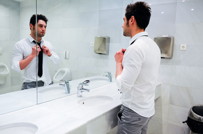 Man adjusting tie in a men's restroom mirror, reflecting discomfort in an unsafe and gross bathroom setting.