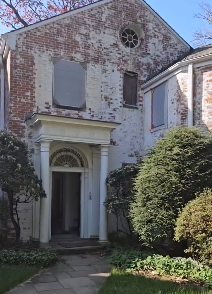 Old brick multimillion-dollar home with boarded windows and classic pillars surrounded by greenery before demolition.